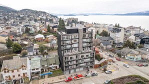 A multi-story building with a modern architectural design, surrounded by a residential area with various houses and a body of water in the background.