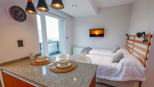 A modern bedroom with a bed, a TV, and a kitchen area with a clock and a dining setup.