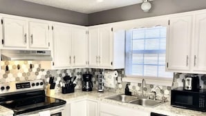 A modern kitchen with white cabinets, stainless steel appliances, a hexagonal tile backsplash, and a window with blinds.