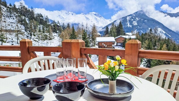 A table with four bowls, a vase with flowers, and a glass set on a deck with a mountain view. - Les Combins 30 by Interhome (Champex)