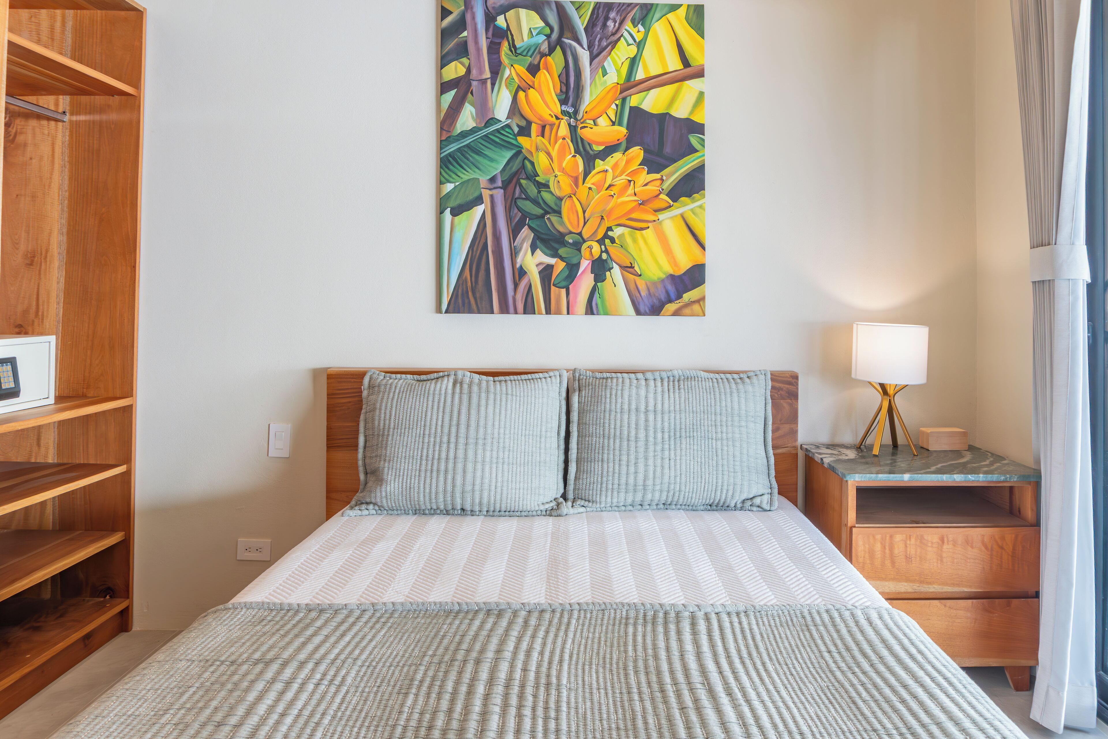 A neatly arranged hotel room with a bed, two pillows, a nightstand with a lamp, and a wall-mounted painting of tropical flowers.