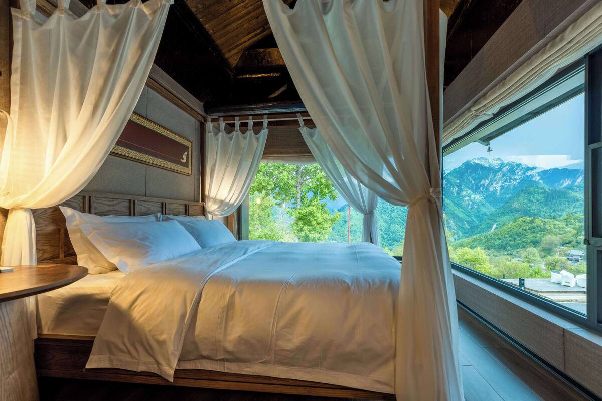 Ein Bett mit weißer Bettwäsche, einem Holz-Kopfteil und transparenten Vorhängen. Ein Fenster bietet Blick auf Berge und Grünflächen.