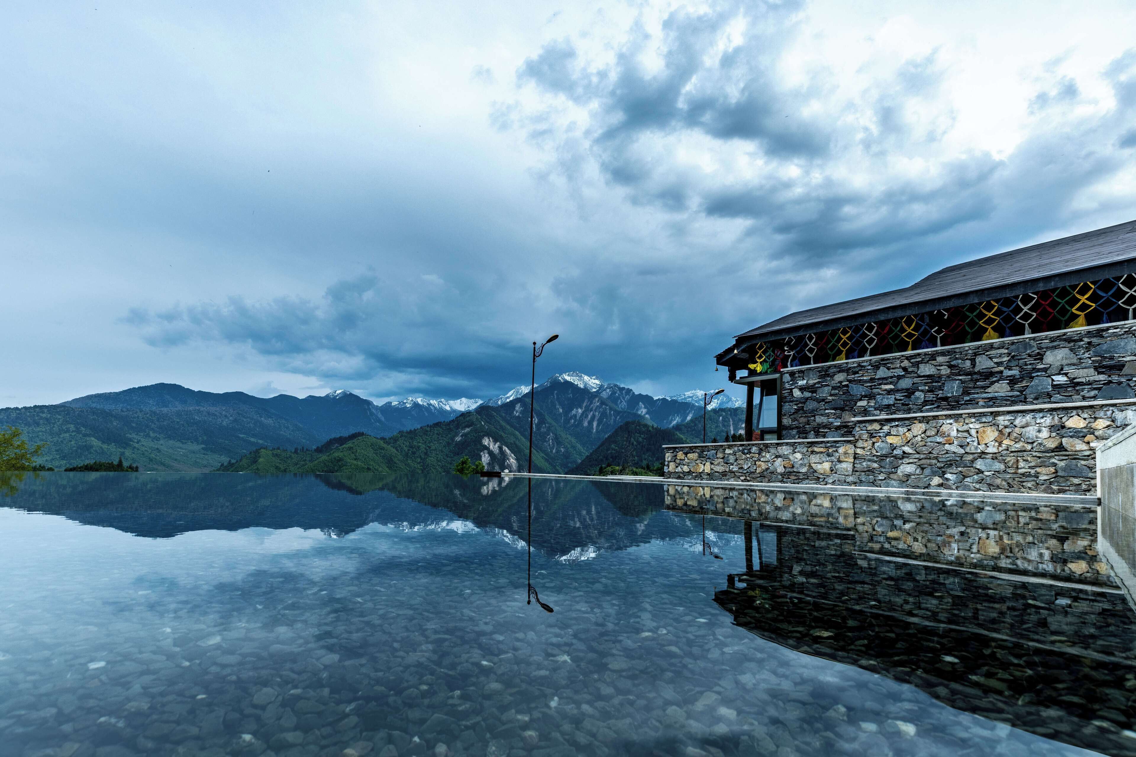 Un edificio in pietra con un riflesso nitido in un lago calmo, circondato da montagne e un cielo nuvoloso.