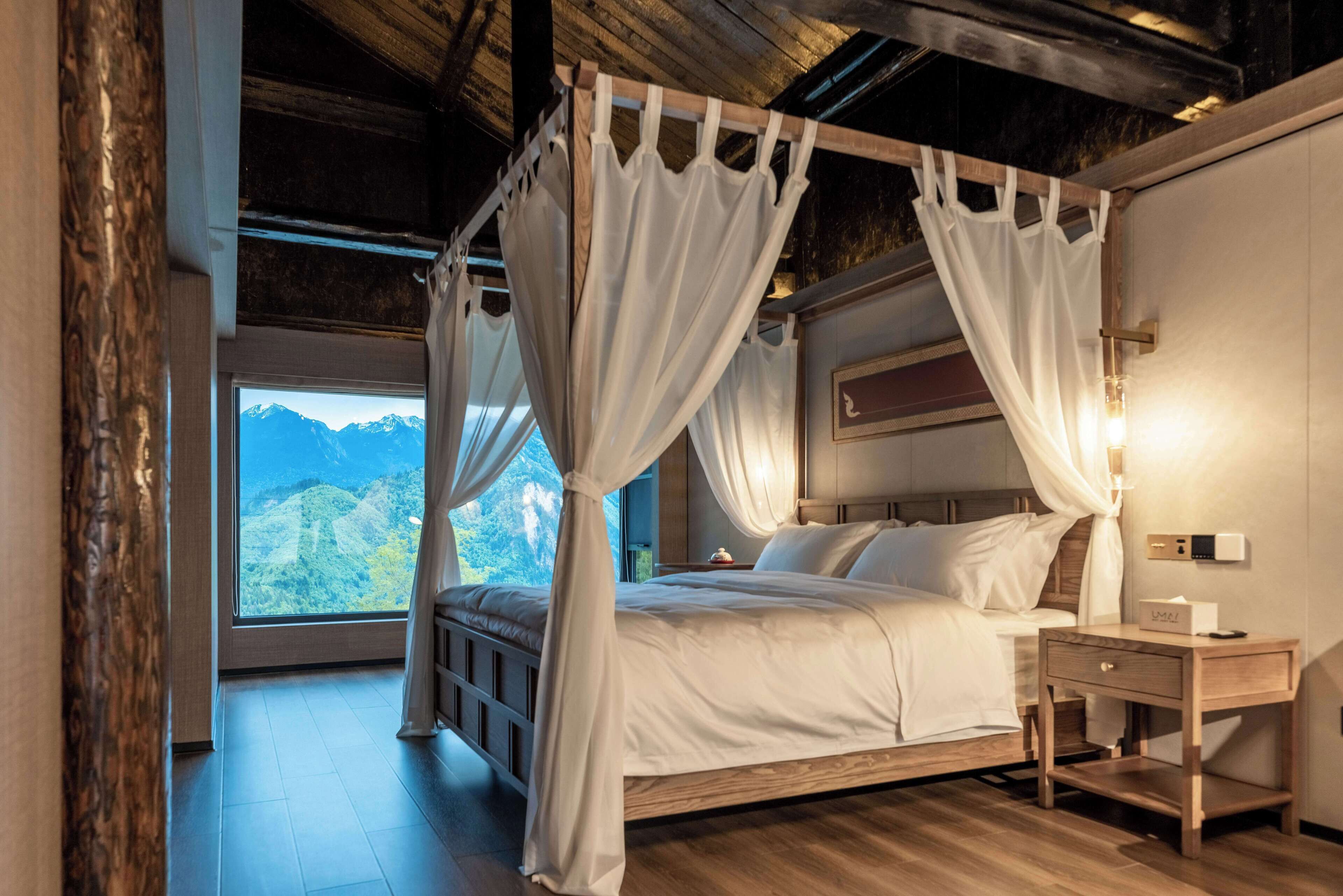 Un lit à baldaquin avec du linge de lit blanc, une table de chevet avec une lampe et une vue sur les montagnes par une grande fenêtre.