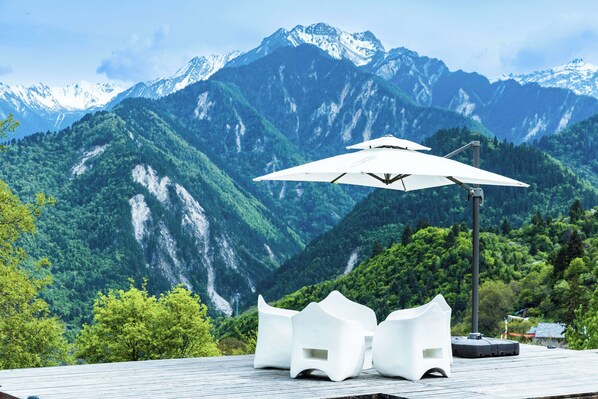 Une terrasse dotée de chaises blanches et d’un parasol blanc, offrant une vue sur un paysage montagneux aux sommets enneigés.