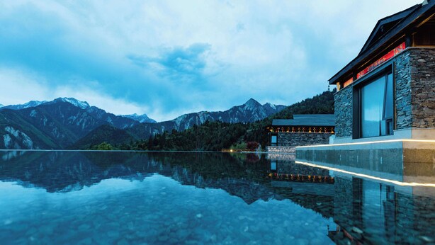 Un edificio moderno con paredes de piedra y ventanas amplias, reflejado en una masa de agua tranquila, rodeado de montañas.