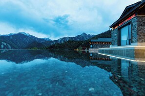 Un bâtiment moderne aux murs de pierre et aux grandes fenêtres, dont le reflet se mire dans une étendue d’eau calme, le tout étant entouré de montagnes.