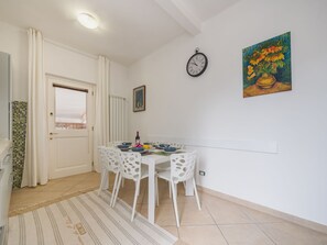 A dining area with a table set for four, white chairs, a clock, and a painting on the wall. - Appartamento La Conchiglia by Interhome (Viareggio)