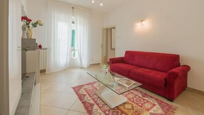 A living room with a red sofa, a glass coffee table, and a patterned rug. There is a white cabinet with a vase of flowers on top.