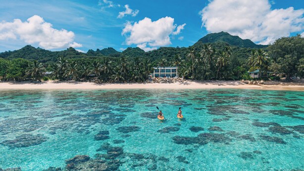 Una playa con agua turquesa cristalina, dos personas haciendo kayak y un edificio de resort con montañas al fondo.