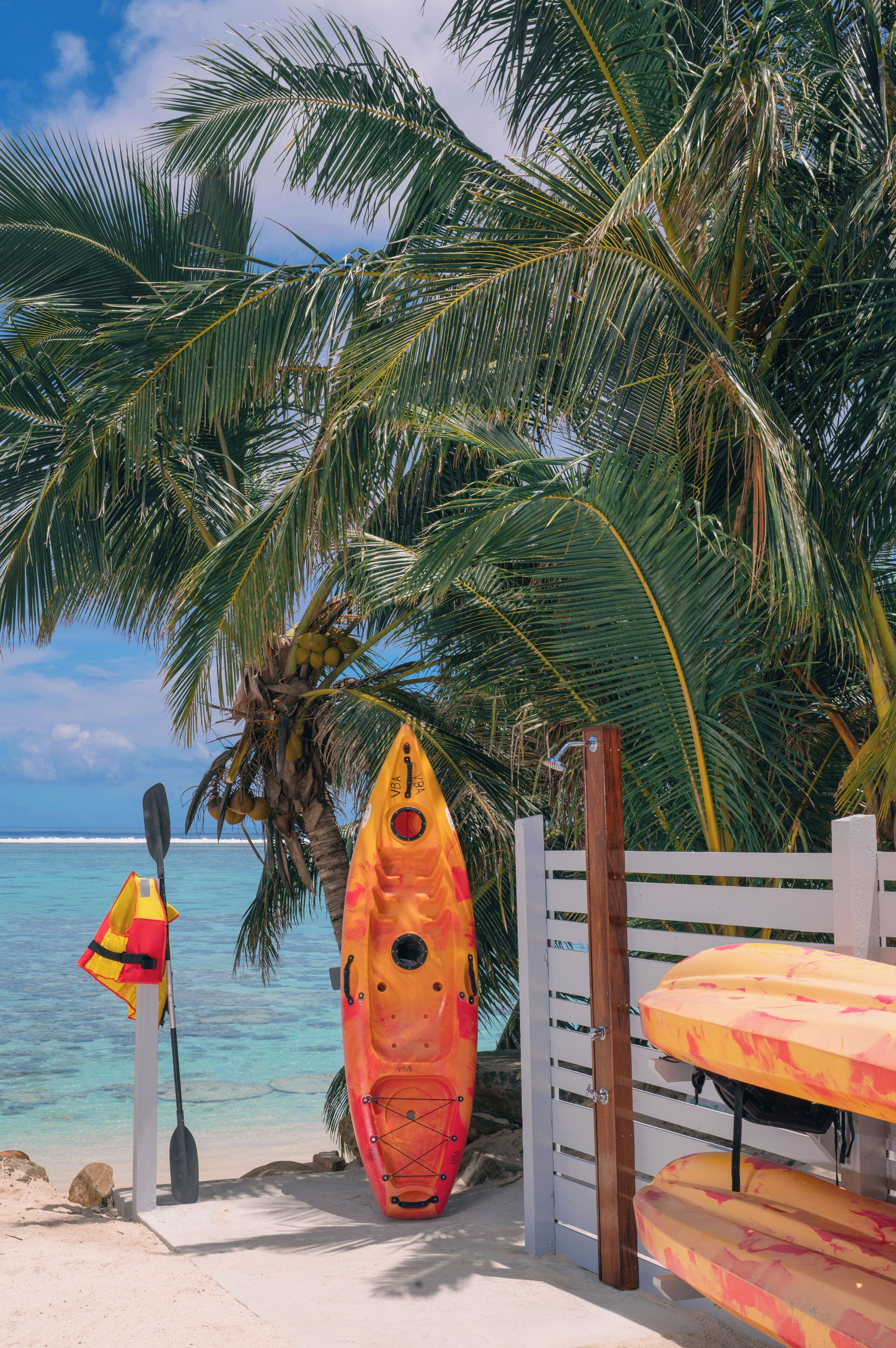Une scène de plage avec un kayak et une pagaie appuyés contre un palmier, une bouée de sauvetage et une clôture en bois.