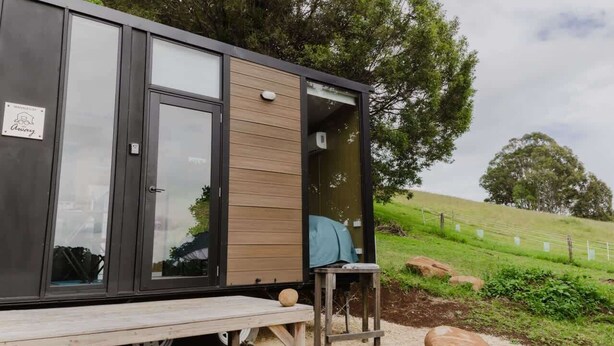 A modern, compact cabin with a sliding door, a small outdoor seating area, and a gravel path.