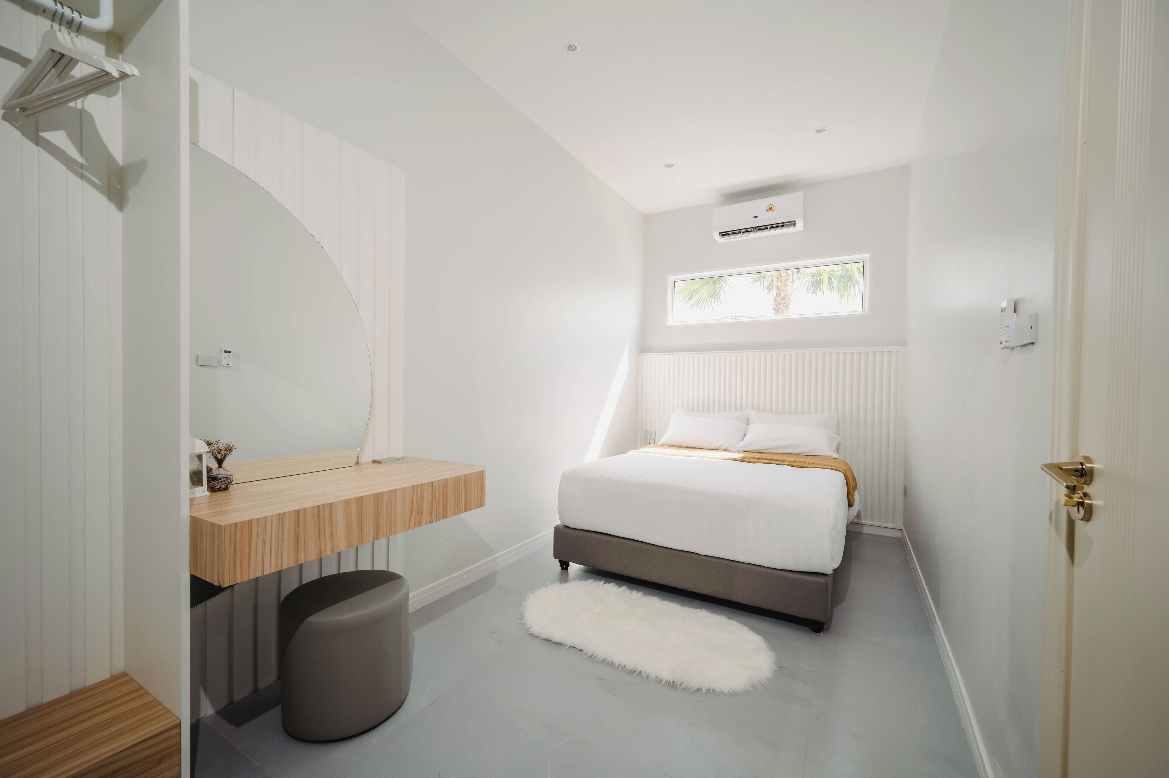 Un dormitorio moderno con una cama, una mesita de noche de madera, un tapete blanco y esponjoso y una ventana con vistas a palmeras.