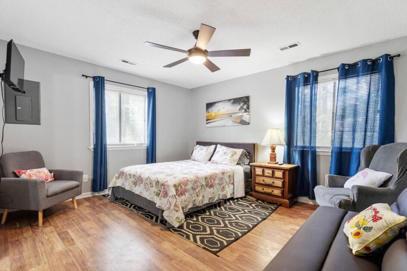 A bedroom with a bed, two chairs, a nightstand, a ceiling fan, and a painting on the wall.