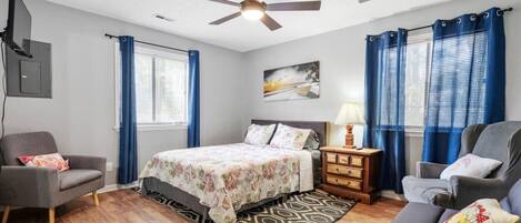 A bedroom with a bed, two chairs, a nightstand, a ceiling fan, and a painting on the wall.