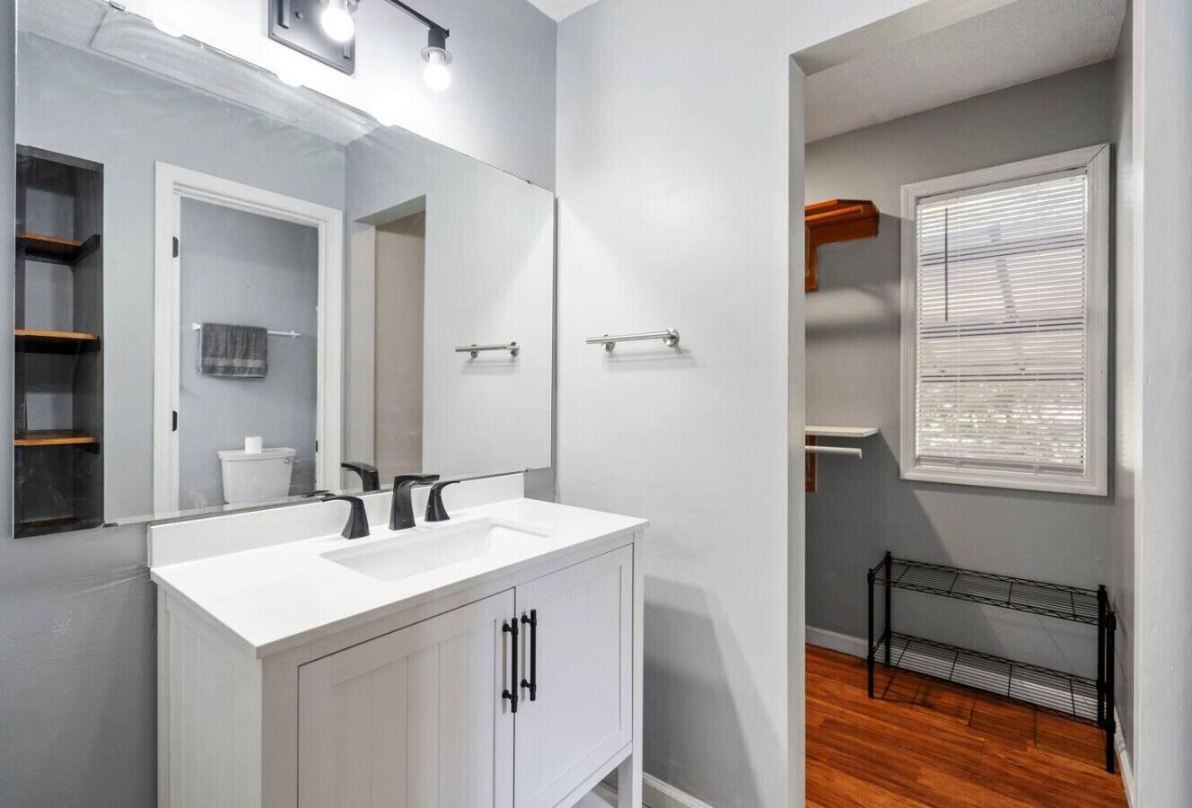 Une salle de bain avec un lavabo blanc, un miroir et des armoires. Il y a une fenêtre avec des stores et une petite étagère sur laquelle est posée une serviette.