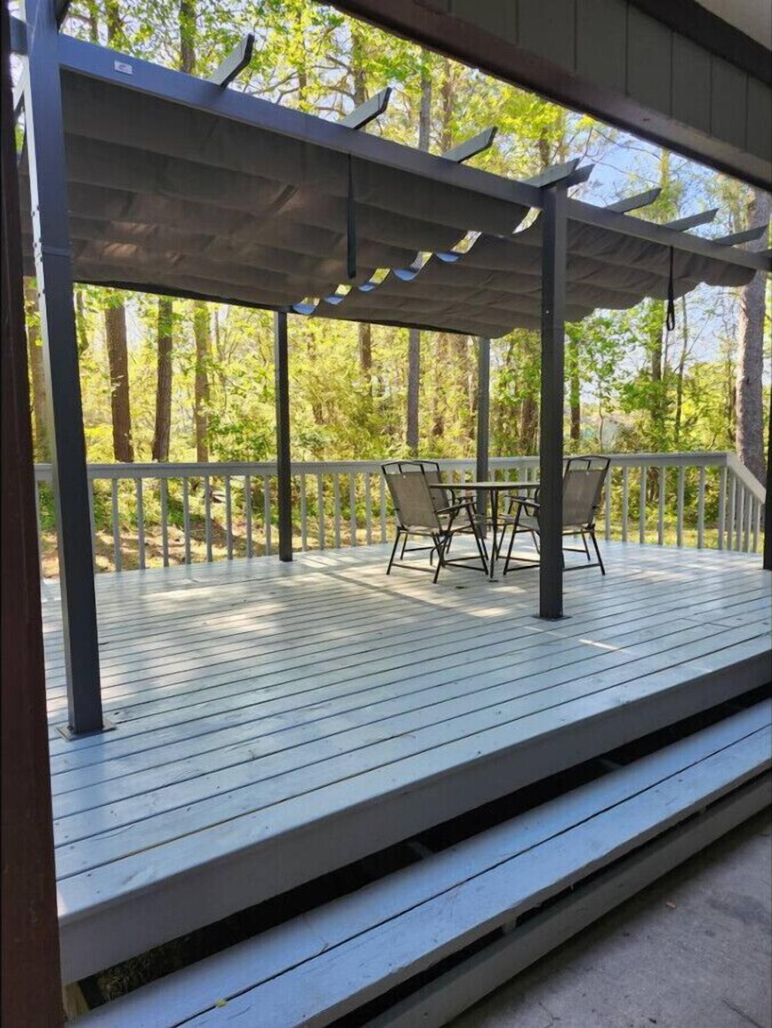A covered outdoor deck with a table and chairs, surrounded by a white railing, set in a wooded area.