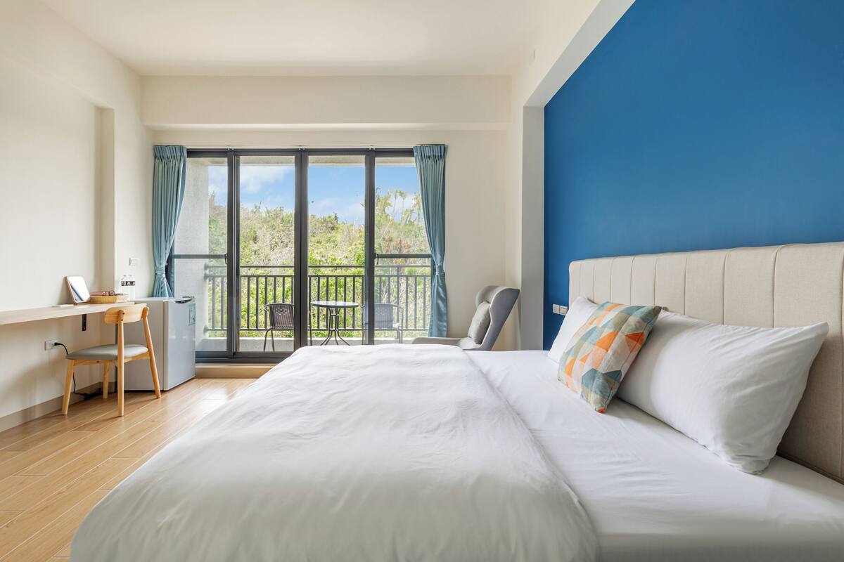 A hotel room with a large bed, a desk, and a chair. There is a balcony with a view of greenery.