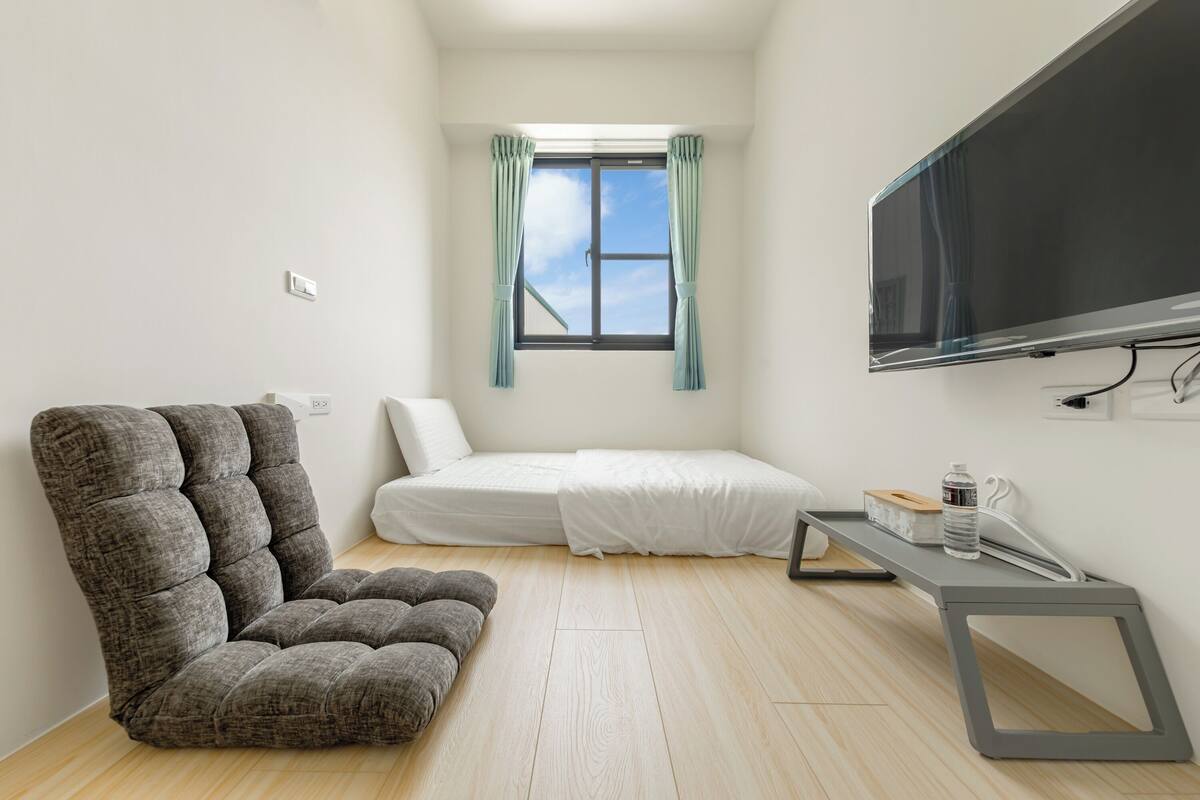 A small, modern room with a sofa, a bed, a TV, and a window with curtains.
