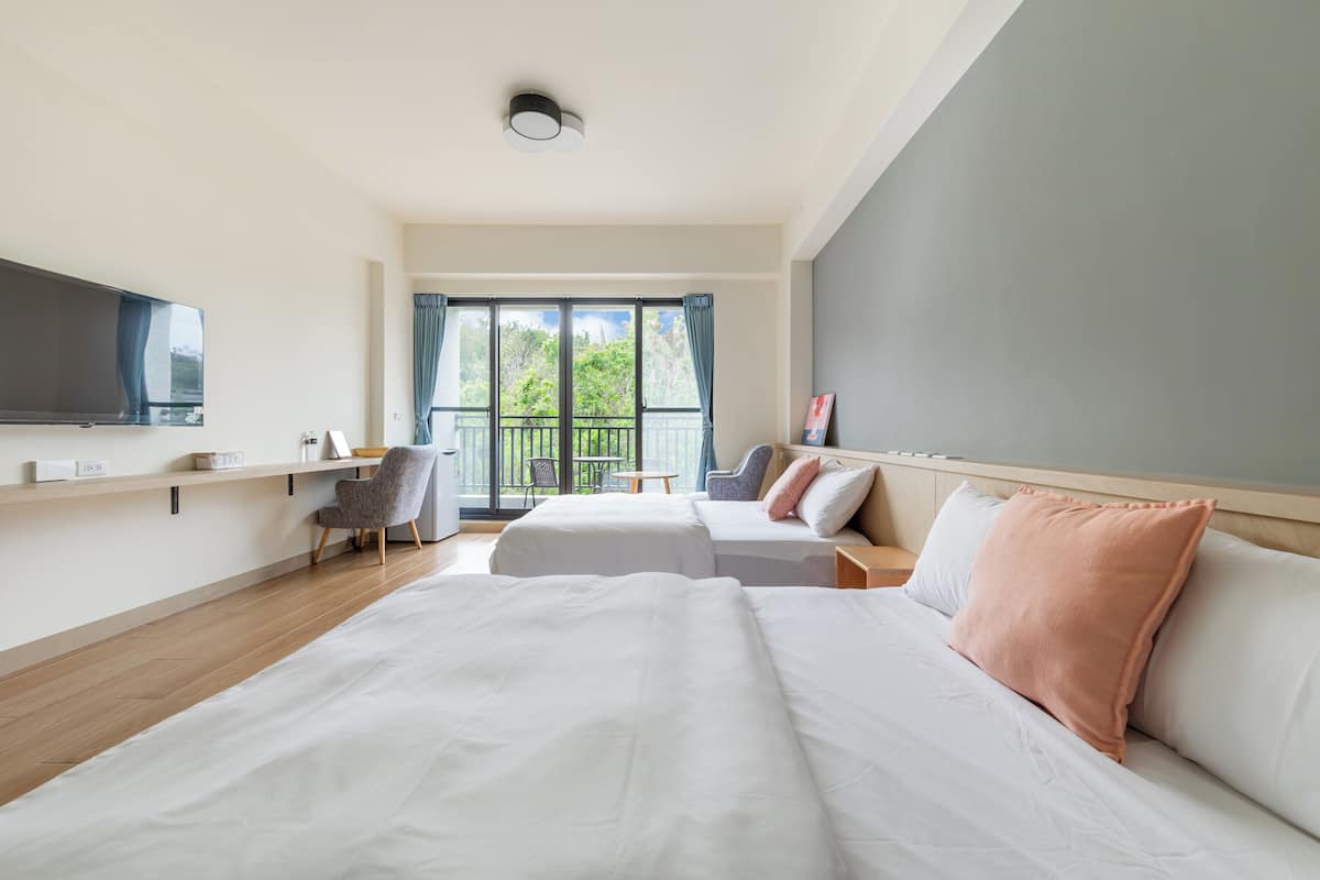 A hotel room with two beds, a TV, a desk, and a balcony with a view of greenery.
