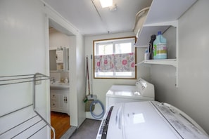A laundry room with a washing machine, a broom, a mop, and a window with floral curtains. - Lulu Manza (Onna)