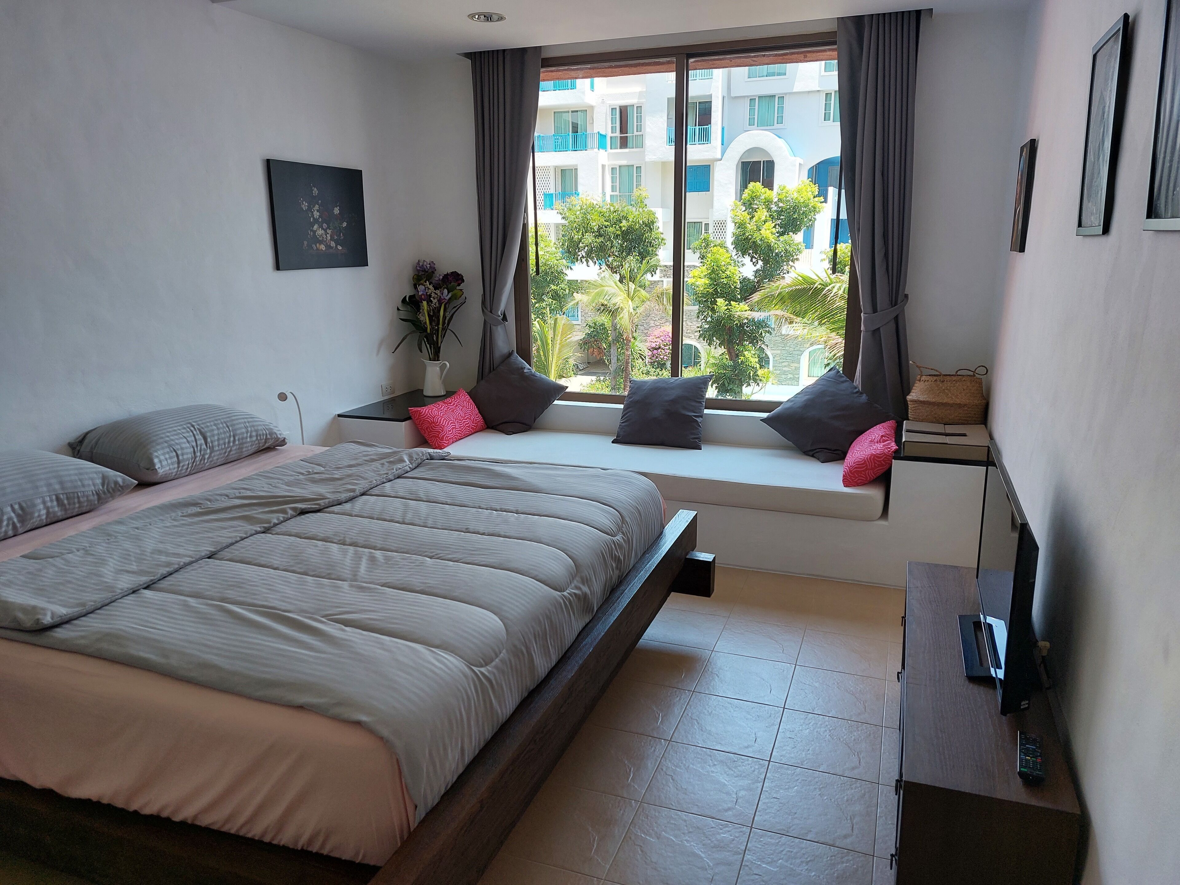 A bedroom with a bed, a window seat with cushions, a bedside table, and a view of buildings outside.