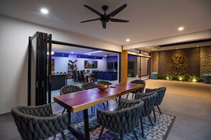 A modern dining area with a wooden table and upholstered chairs, a ceiling fan, and a view into a living space with a blue sofa and artwork. - Stunning 4 bedrooms pool villa at Rawai (Tambon Rawai)