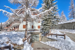 A snow-covered building with a wooden bridge, a stone pathway, and snow-laden trees. - Appartamento Il Rio - 8min Walk to Sportinia Lift (Sauze d'Oulx)