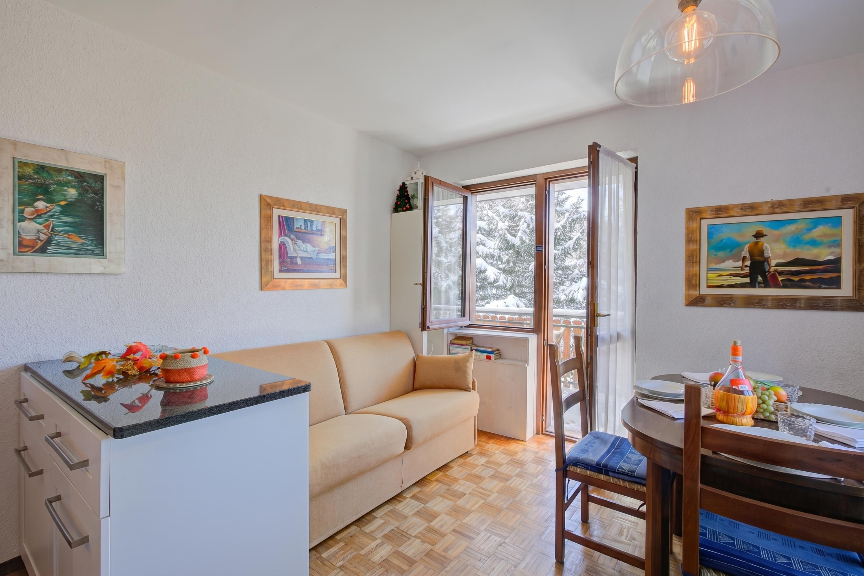 A cozy living area with a sofa, a dining table with chairs, a kitchen area, and a window with a view of snow-covered trees.