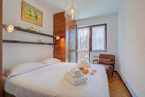 A hotel room with a bed, a chair, a window with a view of trees, and a painting on the wall. - Appartamento Il Rio - 8min Walk to Sportinia Lift (Sauze d'Oulx)