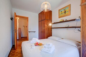Une chambre d’hôtel avec un lit, des serviettes, un tableau, une étagère avec quelques objets et une porte donnant sur une autre pièce.