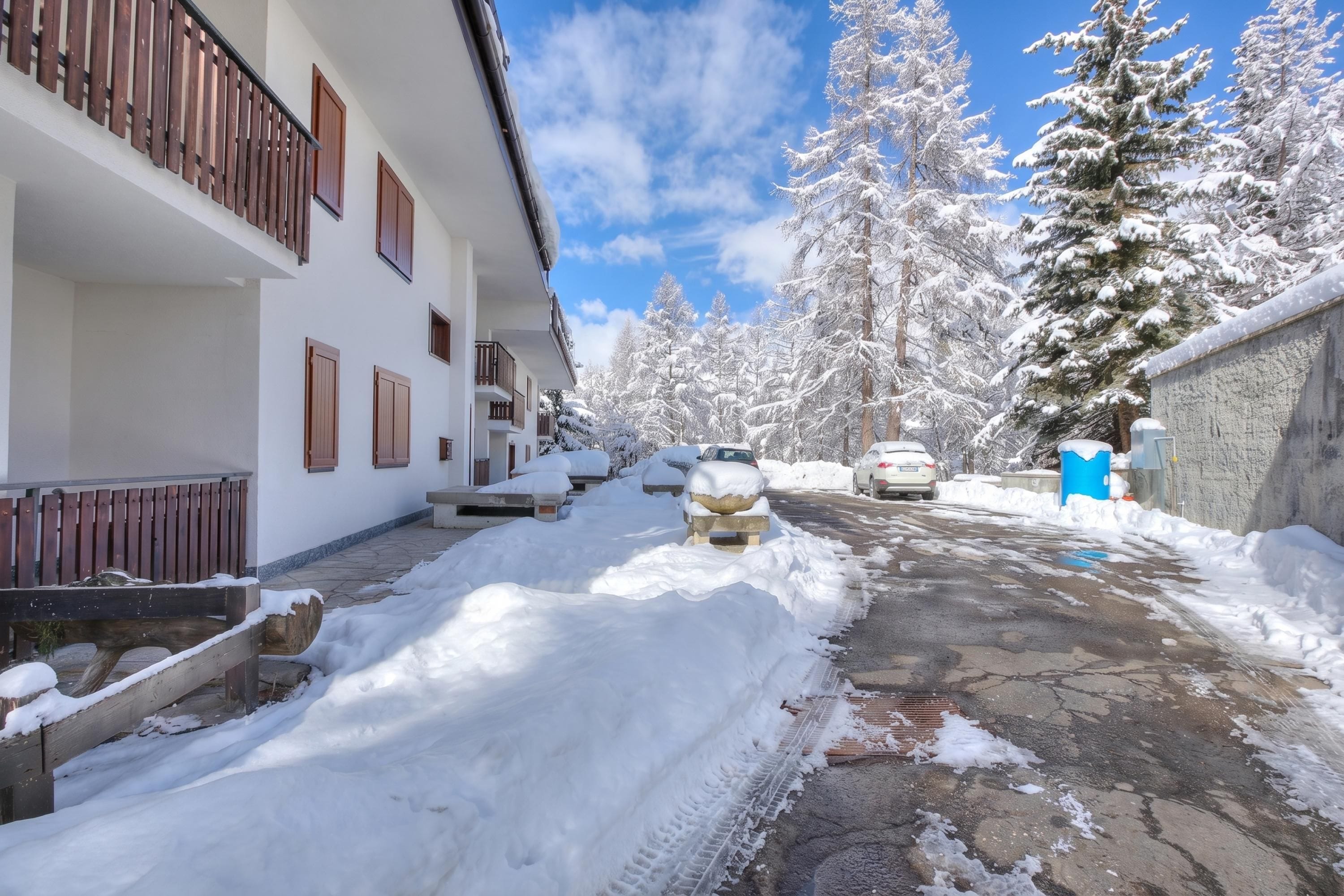 A snow-covered street with a building on the left, a parked car, and snow-laden trees in the background.