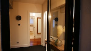 Un pasillo con un reloj en la pared, una puerta que da a otra habitación y un jarrón con plumas sobre una mesa.