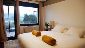 Habitación de hotel con una cama grande, dos toallas enrolladas, una mesita de noche con una lámpara, una ventana con cortinas y vista a zonas verdes.