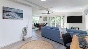 Un salon moderne avec un canapé bleu, un coin repas comprenant une table et des chaises, un grand téléviseur à écran plat et un ventilateur de plafond.