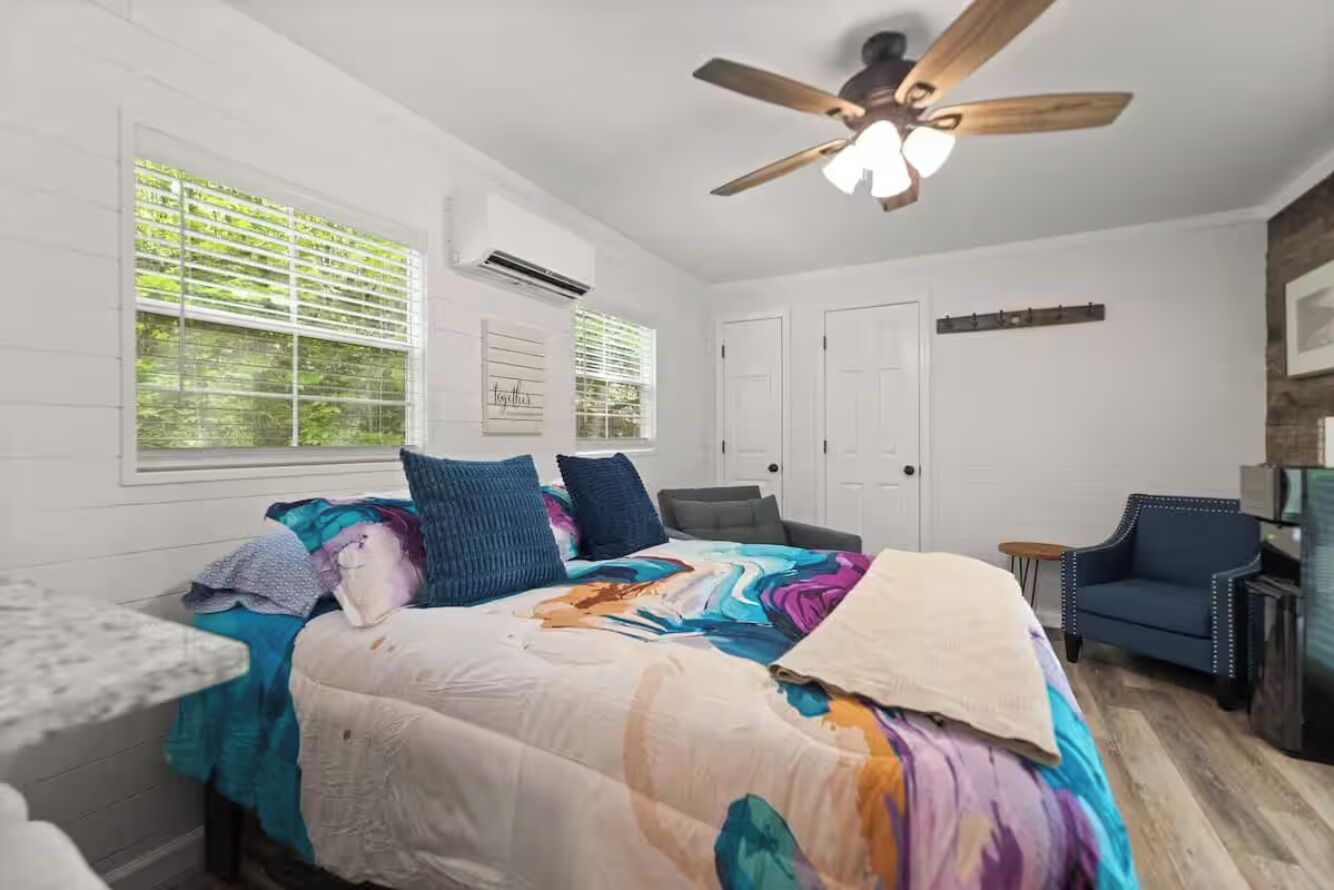 Une chambre à coucher comprenant un lit, un ventilateur de plafond, une fenêtre avec des stores, une porte et une petite table.