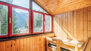 A wooden cabin with a bench and a table, large windows with red frames, and a view of a forested mountain area. - Boiserie by Interhome (Campitello)