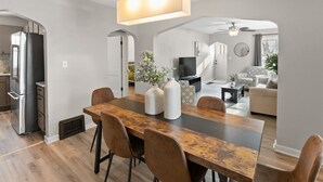 Uma cozinha moderna com geladeira de aço inoxidável, uma área de jantar com mesa e cadeiras de madeira, e uma sala de estar com sofá e televisão.