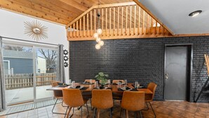 Een eethoek met een houten tafel voor zes personen, omringd door oranje stoelen. Een zwarte bakstenen muur en een houten plafond met zichtbare balken zijn te zien.