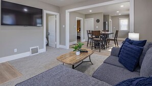 Ein modernes Wohnzimmer mit Flachbildfernseher, Couchtisch aus Holz, grauem Sofa und Essbereich mit Stühlen und Tisch.