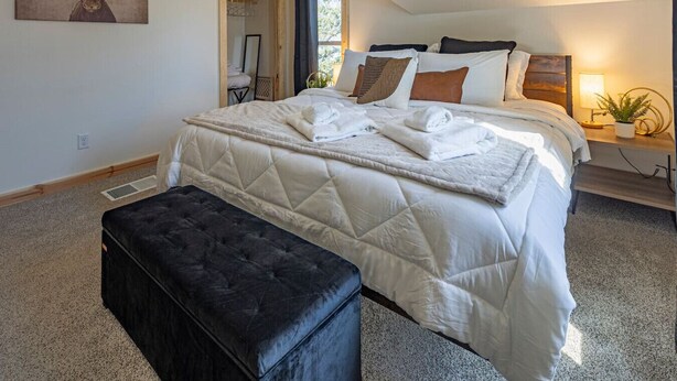 A bedroom with a large bed, a black ottoman, a wooden headboard, a nightstand with a lamp, and a wall-mounted painting of a moose.