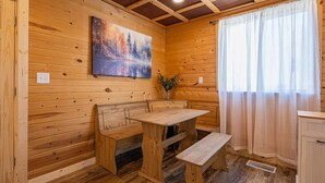 A wooden cabin interior with a wooden table and benches, a painting on the wall, and a window with curtains.