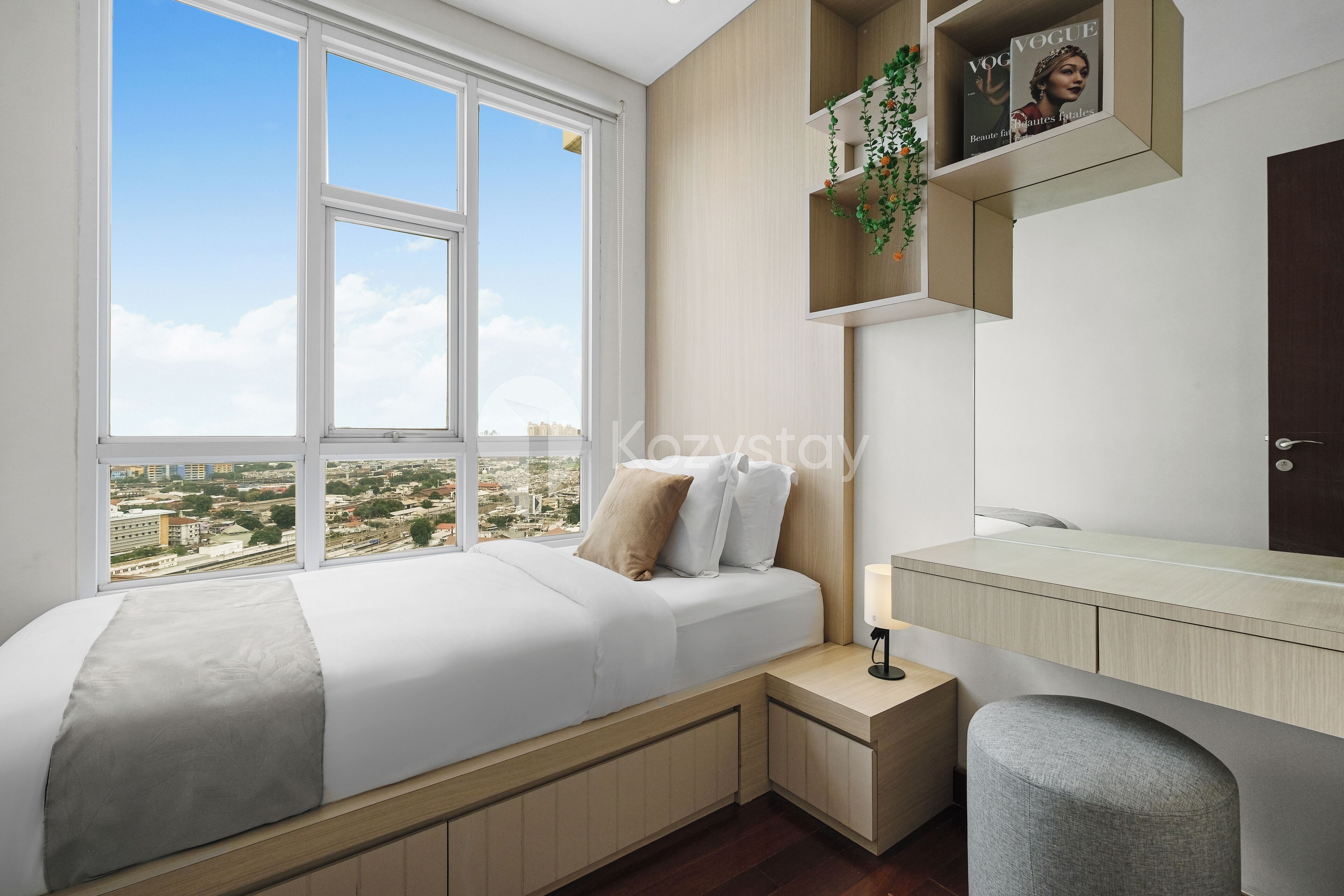 A modern bedroom with a large window, a bed with a grey blanket, a desk, and a shelf with a magazine.