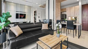 A modern living area with a black leather sofa, a wooden coffee table, and a dining table with chairs. A large mirror reflects the room, and a TV screen displays the word "NETFLIX". - Tensia by Kozystay - Taman Sari (Jakarta)