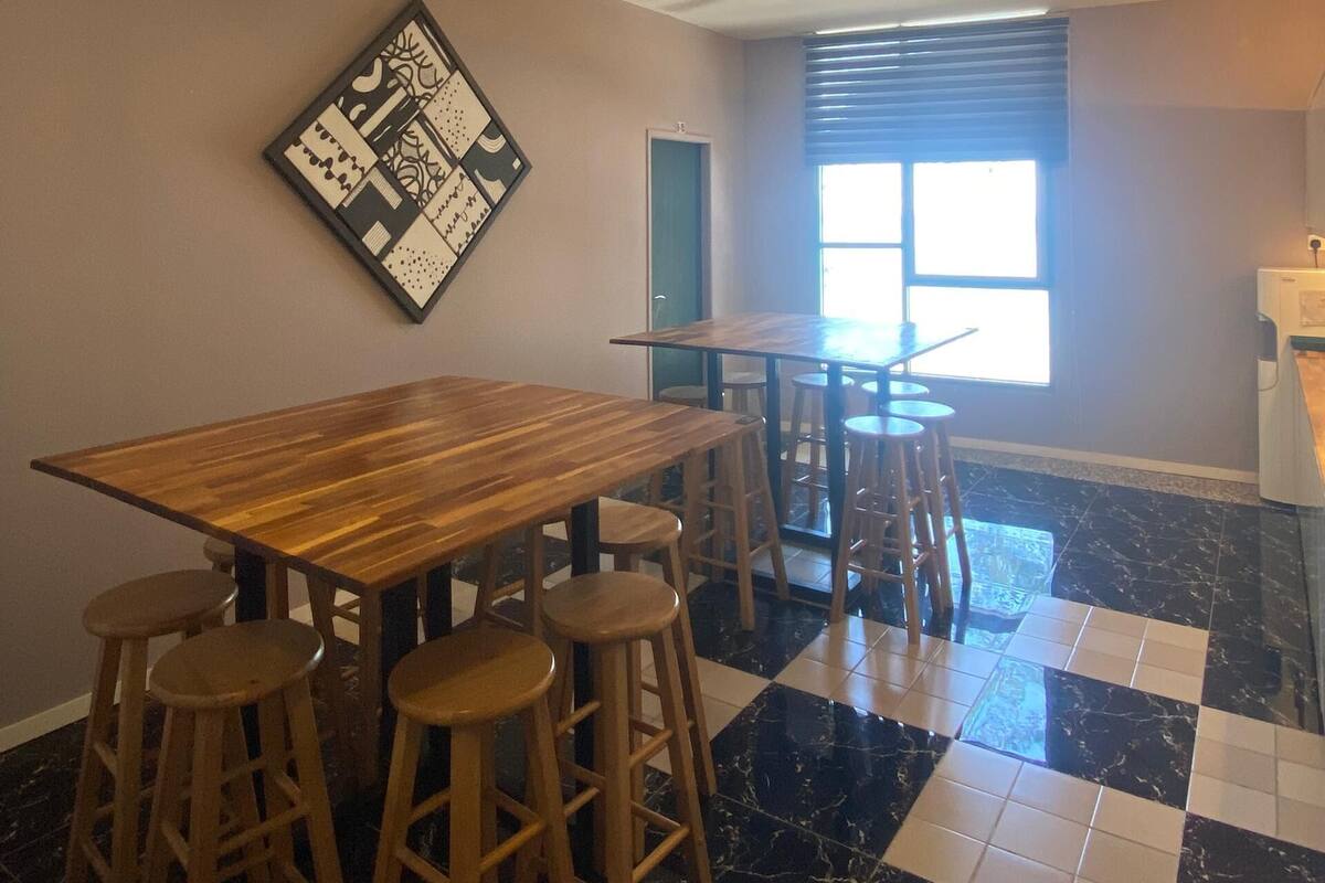 A dining area with a wooden table and bar stools, a ceiling fan, and a window with blinds.