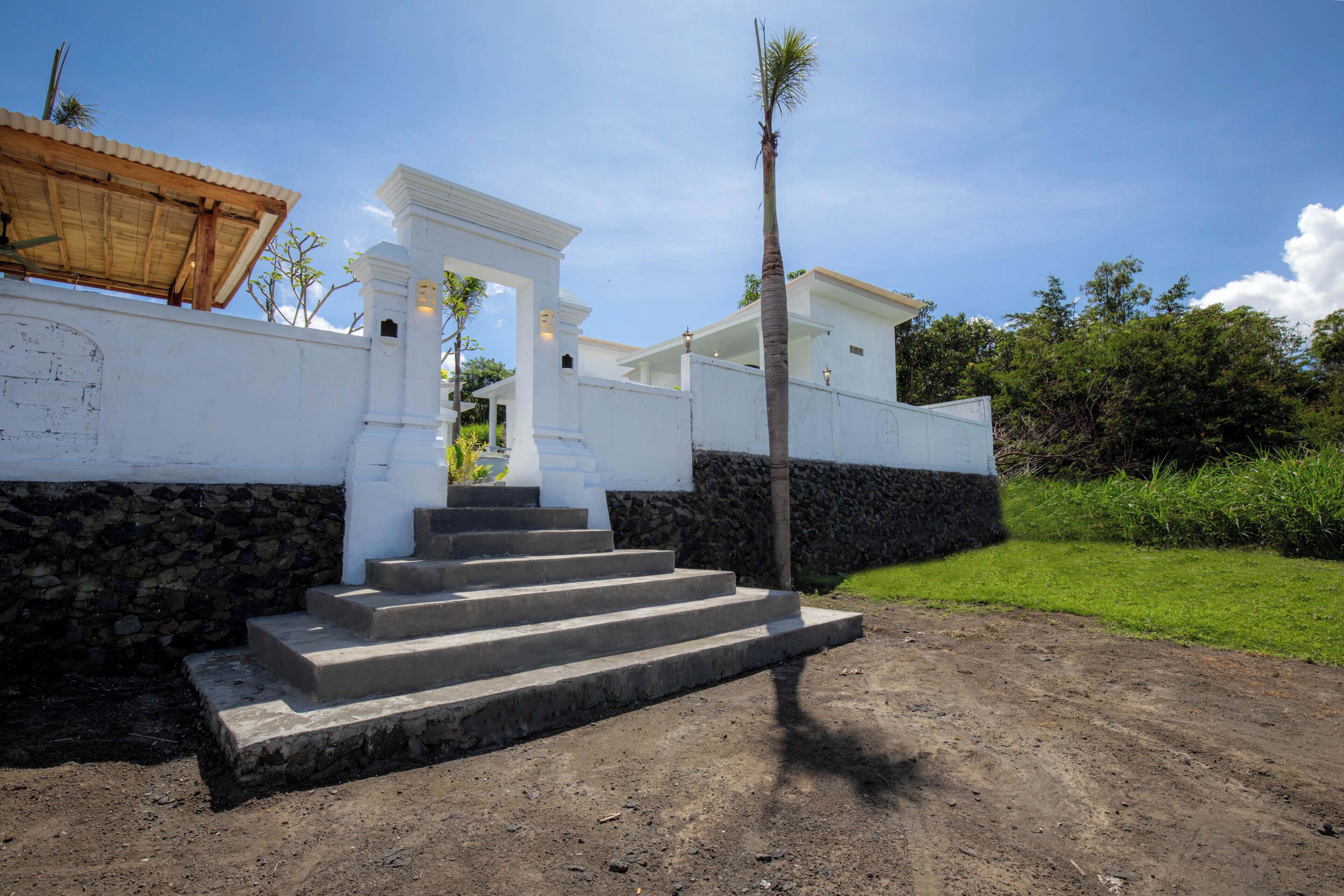 A white building with a stone base, a set of concrete steps leading to a covered entrance, and a palm tree in the foreground.