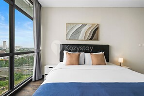 A hotel room with a large window, a bed with white and brown bedding, a nightstand with a lamp, and a city view.