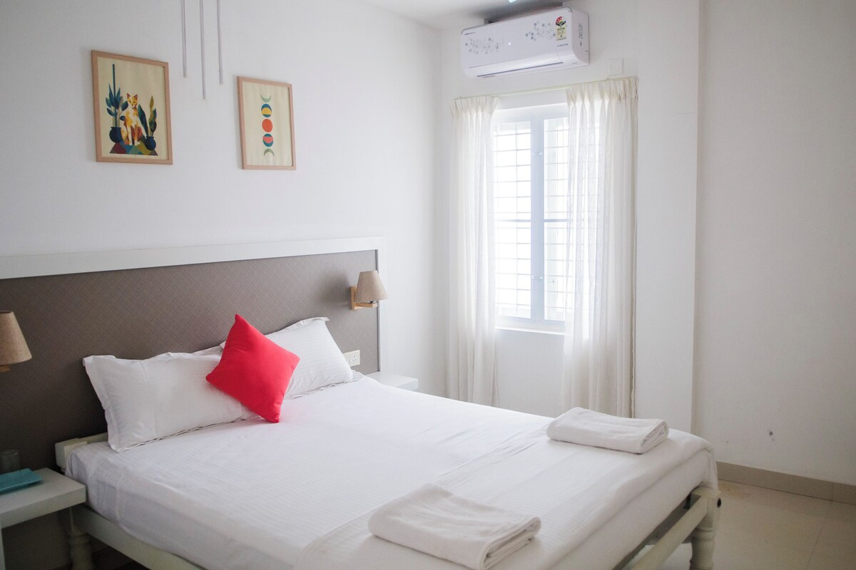 A hotel room with a bed, white bedding, a red pillow, a nightstand, a lamp, and two wall-mounted paintings.