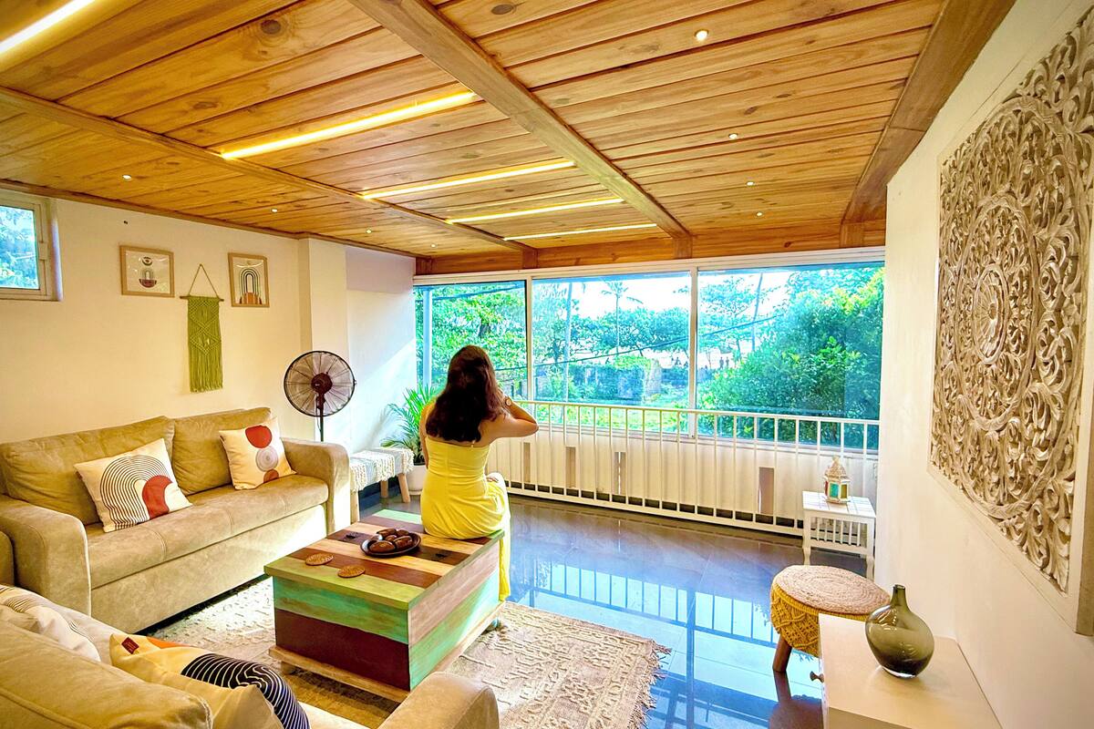 Un soggiorno con soffitto in legno, un divano, un tavolino da caffè e una grande finestra con vista su un’area verde.