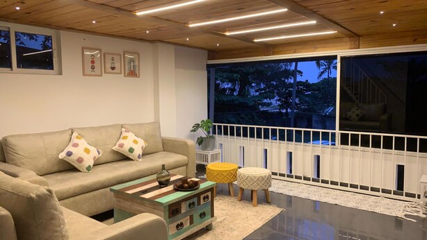 A modern living room with a beige sofa, colorful pillows, a wooden coffee table, and a large window with a view of trees. - Pine Beach House (Alappuzha)